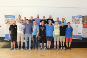 Eine Gruppe von männlichen Personen steht als Gruppe in zwei Reihen hintereinander. Die Personen haben ihre Arme über die Schultern ihrer jeweiligen Nachbarn gelegt.